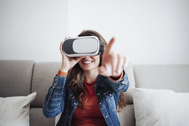 Smiling young woman wearing virtual reality headset and gesturing on sofa - EBBF04897