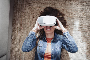 Lächelnde Frau mit Virtual-Reality-Headset auf einem Teppich liegend - EBBF04898