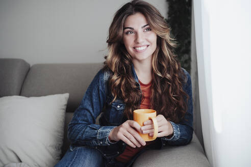 Lächelnde Frau mit Kaffeetasse auf dem Sofa am Fenster sitzend - EBBF04902