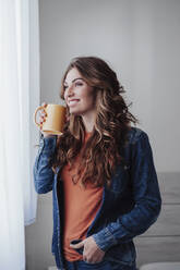 Happy young woman having coffee at window - EBBF04912