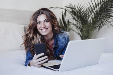 Smiling woman with laptop holding mobile phone on bed - EBBF04916