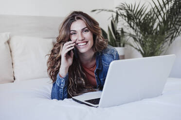 Happy beautiful woman talking on mobile phone in front of laptop at home - EBBF04917