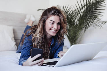 Smiling young woman with long hair using laptop on bed - EBBF04920