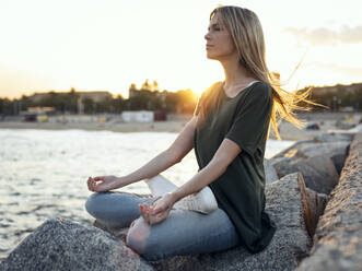 Blond woman meditating on rock at Bogatell beach, Barcelona, Catalonia, Spain - JSRF01713