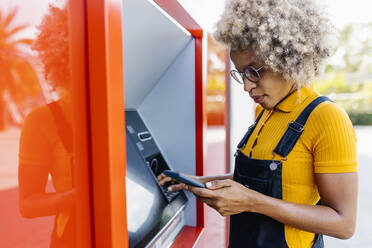 Woman using mobile phone at ATM machine - XLGF02403