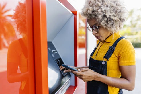 Frau benutzt Mobiltelefon am Geldautomaten - XLGF02403