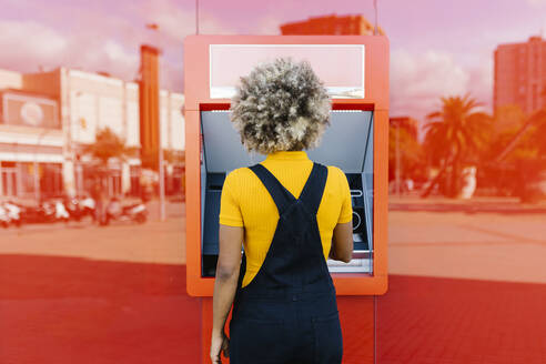 Woman with afro hairstyle using red ATM machine - XLGF02404