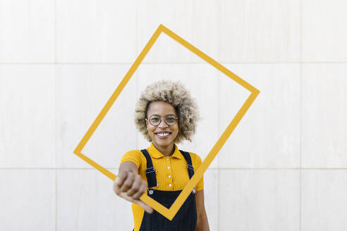 Frau hält gelben Rahmen vor einer Wand - XLGF02433