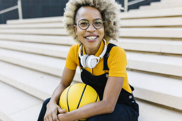 Lächelnde Frau mit Brille sitzt mit Basketball auf den Stufen - XLGF02467