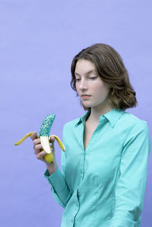 Ernstes Mädchen hält glitzernde Banane vor lavendelfarbenem Hintergrund - PSTF00990