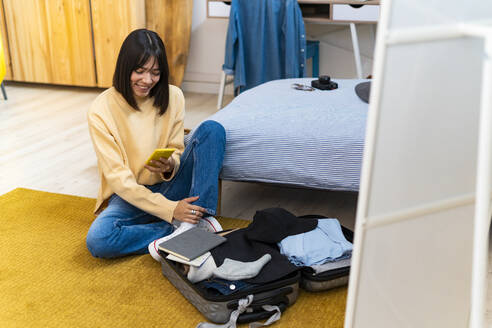 Lächelnde junge Frau mit Smartphone packt Tasche im Schlafzimmer - GIOF14224