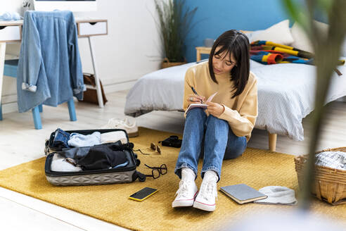 Junge Frau schreibt in Tagebuch, während sie im Schlafzimmer ihre Tasche packt - GIOF14232