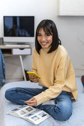 Lächelnde junge Frau mit Foto und Smartphone auf dem Bett zu Hause - GIOF14240
