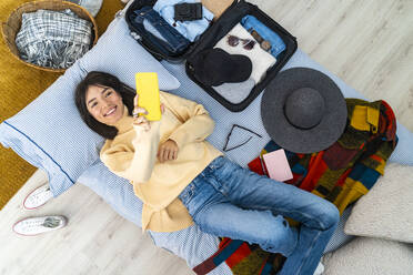 Smiling woman taking selfie with smart phone while lying by luggage on bed - GIOF14242