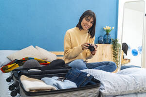 Junge Frau mit Kamera sitzt in der Nähe von Gepäck auf dem Bett zu Hause - GIOF14245
