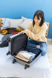 Lächelnde junge Frau packt Koffer auf dem Bett zu Hause - GIOF14255