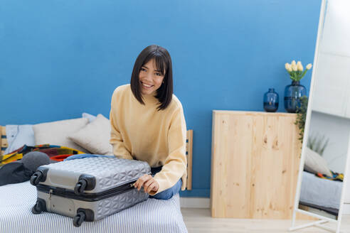 Glückliche junge Frau packt Tasche auf dem Bett zu Hause - GIOF14256