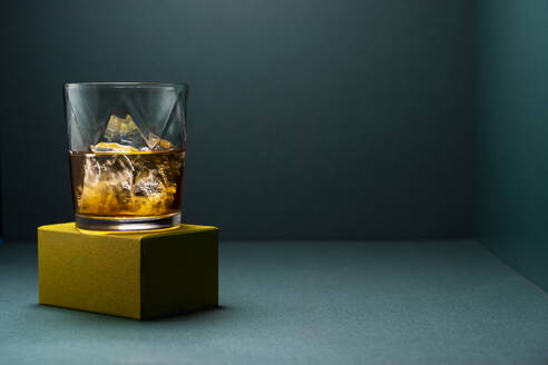 Glasbecher mit kaltem Whiskey und Eiswürfeln auf einem goldfarbenen zerklüfteten Boden und einem türkisblauen Eckhintergrund - ADSF31816
