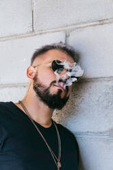 Thoughtful male with beard and mustache and with smoke coming out of his mouth in casual clothes and sunglasses standing near grey wall of building - ADSF31825