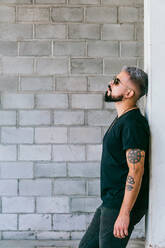 Side view of pensive male with beard and tattoos on arms in black outfit and sunglasses standing and leaning on grey wall of building in daylight - ADSF31826