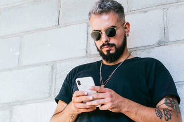 Happy bearded guy with tattoos in black t shirt and sunglasses standing near wall of building and using a smartphone in daylight - ADSF31827