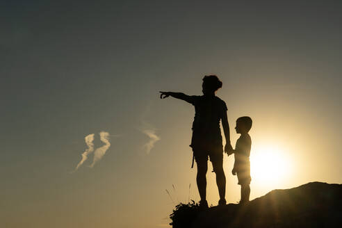 Mother and son against the light silhouetted by the sun pointing with their arms towards the horizon at sunset - ADSF31832