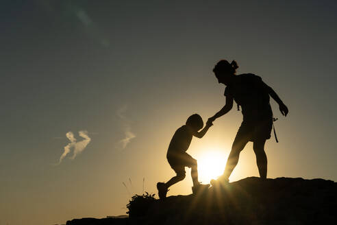 Mother helping her son climb a mountain against the light silhouetted by the sun in a motivational story - ADSF31833