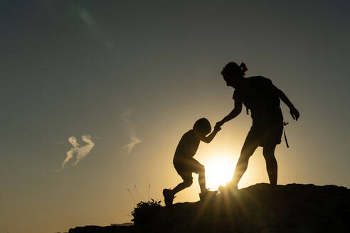 Mutter, die ihrem Sohn beim Besteigen eines Berges im Gegenlicht der Sonne hilft, in einer Motivationsgeschichte - ADSF31833