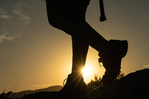 Silhouette der Beine einer Frau beim Trekking in den Bergen mit der Sonne, die einen Sonnenstern mit ihrem Fuß bei Sonnenuntergang erzeugt - ADSF31834