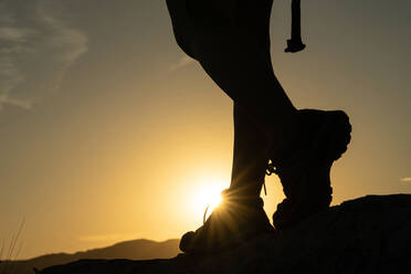 Silhouette der Beine einer Frau beim Trekking in den Bergen mit der Sonne, die einen Sonnenstern mit ihrem Fuß bei Sonnenuntergang erzeugt - ADSF31836