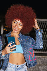 Cheerful female with Afro hairstyle wearing trendy denim outfit text messaging on cellphone while standing near fence on street in evening time - ADSF31844
