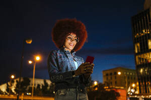 Positive Frau mit Afrofrisur und moderner Kleidung, die eine Textnachricht auf dem Handy schreibt, während sie auf einer Straße mit Gebäuden und Straßenlaternen in der Abendzeit steht - ADSF31845
