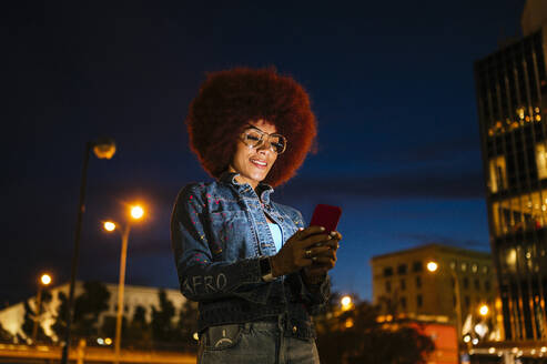 Positive female with Afro hairstyle and modern clothes text messaging on cellphone while standing on street with buildings and streetlamps in evening time - ADSF31845