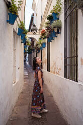 Full body side view of smiling Asian female tourist standing in narrow passage between houses while observing flowerpots in town - ADSF31851