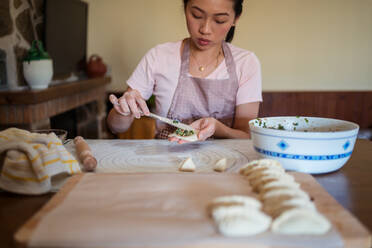 Frau in Freizeitkleidung und Schürze füllt Teigtaschen mit Fleisch, während sie in der Küche traditionelle chinesische Jiaozi zubereitet - ADSF31860