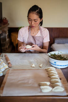 Frau in Freizeitkleidung und Schürze füllt Teigtaschen mit Fleisch, während sie in der Küche traditionelle chinesische Jiaozi zubereitet - ADSF31861