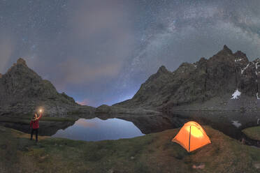 Picturesque scenery of unrecognizable traveler standing with bright light in hand near tent placed among rocky mountains under cloudless night sky milky way located in Circo de Gredos cirque in Spain - ADSF31872