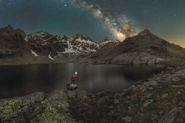 Back view of unrecognizable male tourist with torch admiring snowy mounts reflecting in water under starry sky at night - ADSF31873