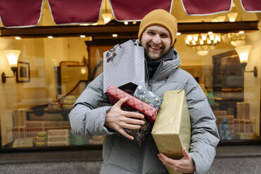 Smiling man holding Christmas presents in front of store - EYAF01809