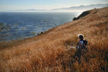 Eine junge Wanderin ruht sich in einem wilden Grasfeld an der Küste der neuseeländischen Halbinsel Kaikoura aus, Region Canterbury, Südinsel, Neuseeland, Pazifik - RHPLF20879