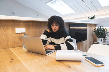 Curly haired businesswoman working on laptop in office - JAQF00984