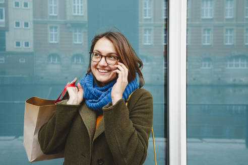 Lächelnde Frau mit Einkaufstasche, die vor einer Glaswand mit einem Smartphone telefoniert - IHF00601