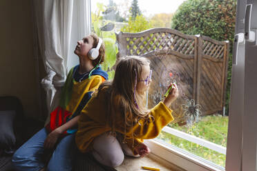 Mädchen hört Musik, während ihre Schwester eine Zeichnung auf einem transparenten Fenster anfertigt - IHF00615