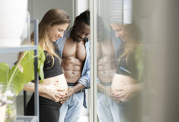 Mann berührt den Bauch einer schwangeren Frau, die zu Hause am Fenster steht - JCCMF04654