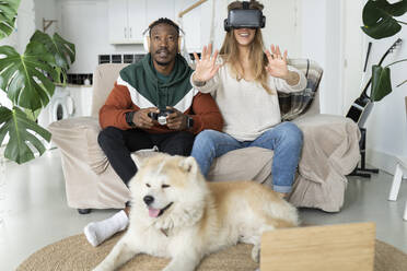Lächelnde Frau, die ein Virtual-Reality-Headset trägt, während ein Mann ein Videospiel vor seinem Hund zu Hause spielt - JCCMF04684