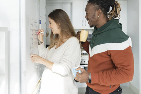 Pregnant woman with man in kitchen crossing date on calendar at home - JCCMF04688