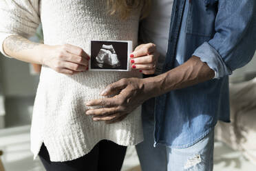 Man touching belly of pregnant woman holding ultrasound scan at home - JCCMF04692