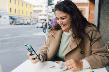 Glückliche Frau, die ihr Smartphone benutzt, während sie in einem Straßencafé Kaffee trinkt - MEUF04797