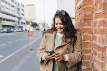 Smiling curvy woman using mobile phone while leaning on brick wall - MEUF04799