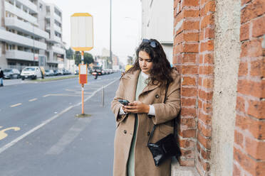 Voluptuous woman using mobile phone while leaning on brick wall - MEUF04804